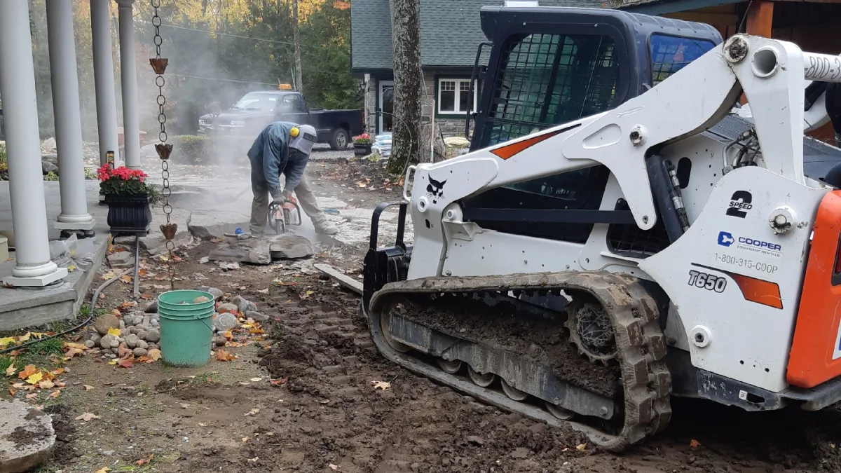 A worker cutting stone near a front porch with a tracked loader alongside